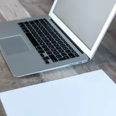 Laptop z otwartym, białym ekranem stoi na drewnianym biurku obok kartki papieru.