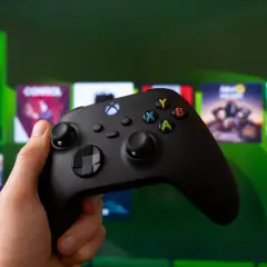 Dłoń trzyma czarny kontroler Xbox, gotowa do gry. Na ekranie w tle widać logo Xbox Game Pass i okładki gier.