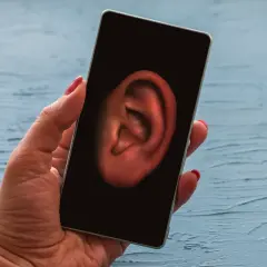 Dłoń trzyma smartfon z uchem na ekranie. Czy Twój telefon jest na podsłuchu? Dowiedz się, jak sprawdzić.
