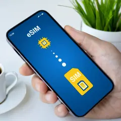 Dłoń trzyma smartfon z ekranem pokazującym grafikę symbolizującą play esim. Obok filiżanka kawy.