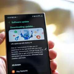Dłoń trzyma smartfon z ekranem pokazującym aktualizację oprogramowania One UI 7. "Downloading update..." informuje o postępie.