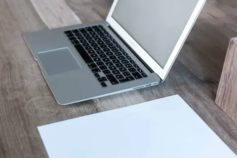 Laptop z otwartym, białym ekranem stoi na drewnianym biurku obok kartki papieru.