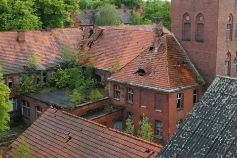Opuszczony **szpital psychiatryczny** z rozpadającymi się dachami i zarośniętymi dziedzińcami, idealne miejsce na **horror o szpitalu psychiatrycznym**.