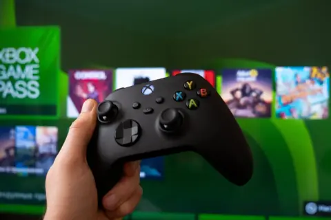 Dłoń trzyma czarny kontroler Xbox, gotowa do gry. Na ekranie w tle widać logo Xbox Game Pass i okładki gier.