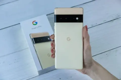 Dłoń trzyma kremowy telefon Pixel 6 Pro, obok pudełko z tymi samymi telefonami z czystym Androidem.