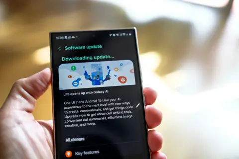 Dłoń trzyma smartfon z ekranem pokazującym aktualizację oprogramowania One UI 7. "Downloading update..." informuje o postępie.