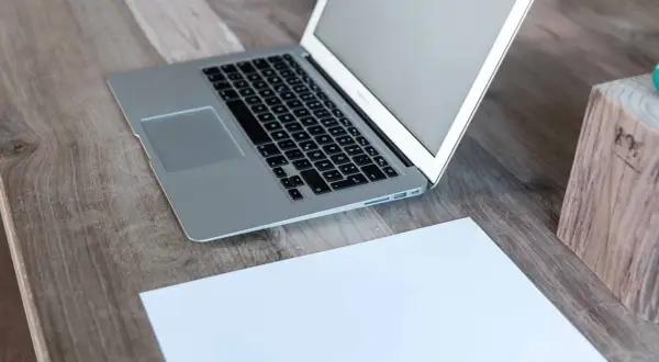 Laptop z otwartym, białym ekranem stoi na drewnianym biurku obok kartki papieru.