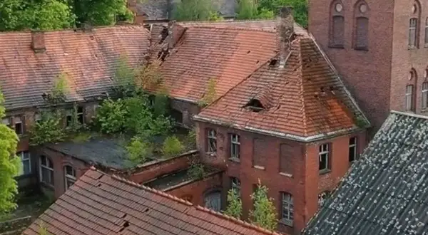 Opuszczony **szpital psychiatryczny** z rozpadającymi się dachami i zarośniętymi dziedzińcami, idealne miejsce na **horror o szpitalu psychiatrycznym**.