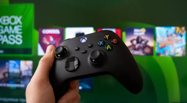 Dłoń trzyma czarny kontroler Xbox, gotowa do gry. Na ekranie w tle widać logo Xbox Game Pass i okładki gier.
