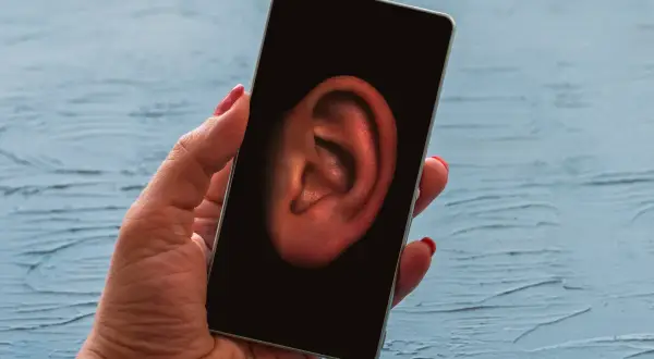 Dłoń trzyma smartfon z uchem na ekranie. Czy Twój telefon jest na podsłuchu? Dowiedz się, jak sprawdzić.