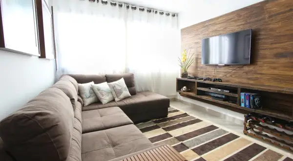 Nowoczesny salon z brązową kanapą, telewizorem na drewnianej ścianie i dywanem. Idealny przykład, jaki uchwyt do telewizora wybrać, by stworzyć taką przestrzeń.