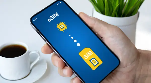 Dłoń trzyma smartfon z ekranem pokazującym grafikę symbolizującą play esim. Obok filiżanka kawy.