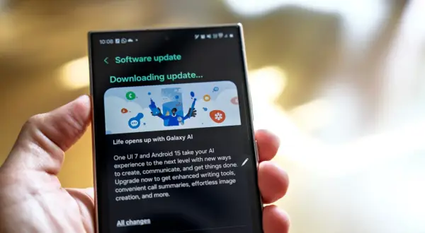 Dłoń trzyma smartfon z ekranem pokazującym aktualizację oprogramowania One UI 7. "Downloading update..." informuje o postępie.