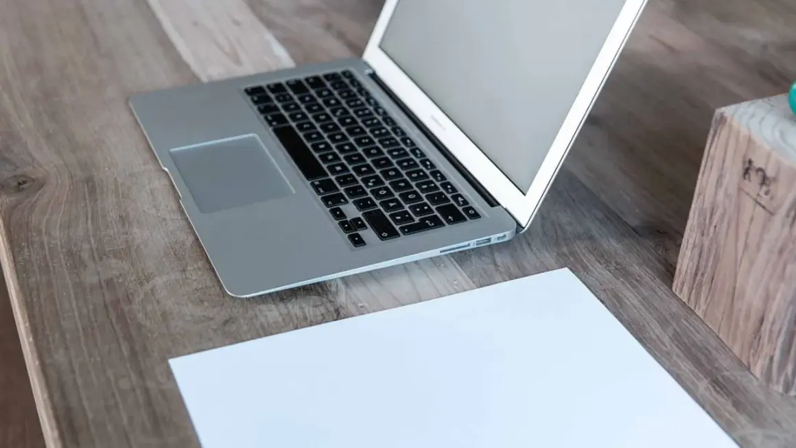 Laptop z otwartym, białym ekranem stoi na drewnianym biurku obok kartki papieru.