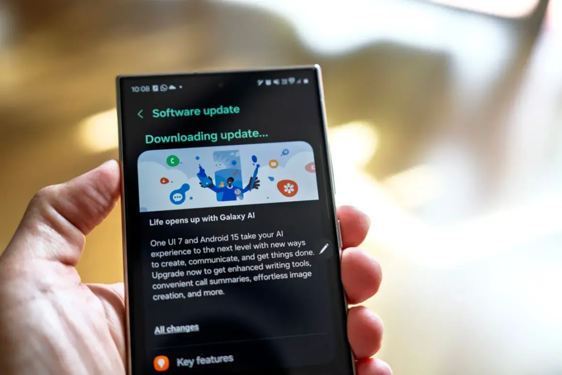 Dłoń trzyma smartfon z ekranem pokazującym aktualizację oprogramowania One UI 7. "Downloading update..." informuje o postępie.