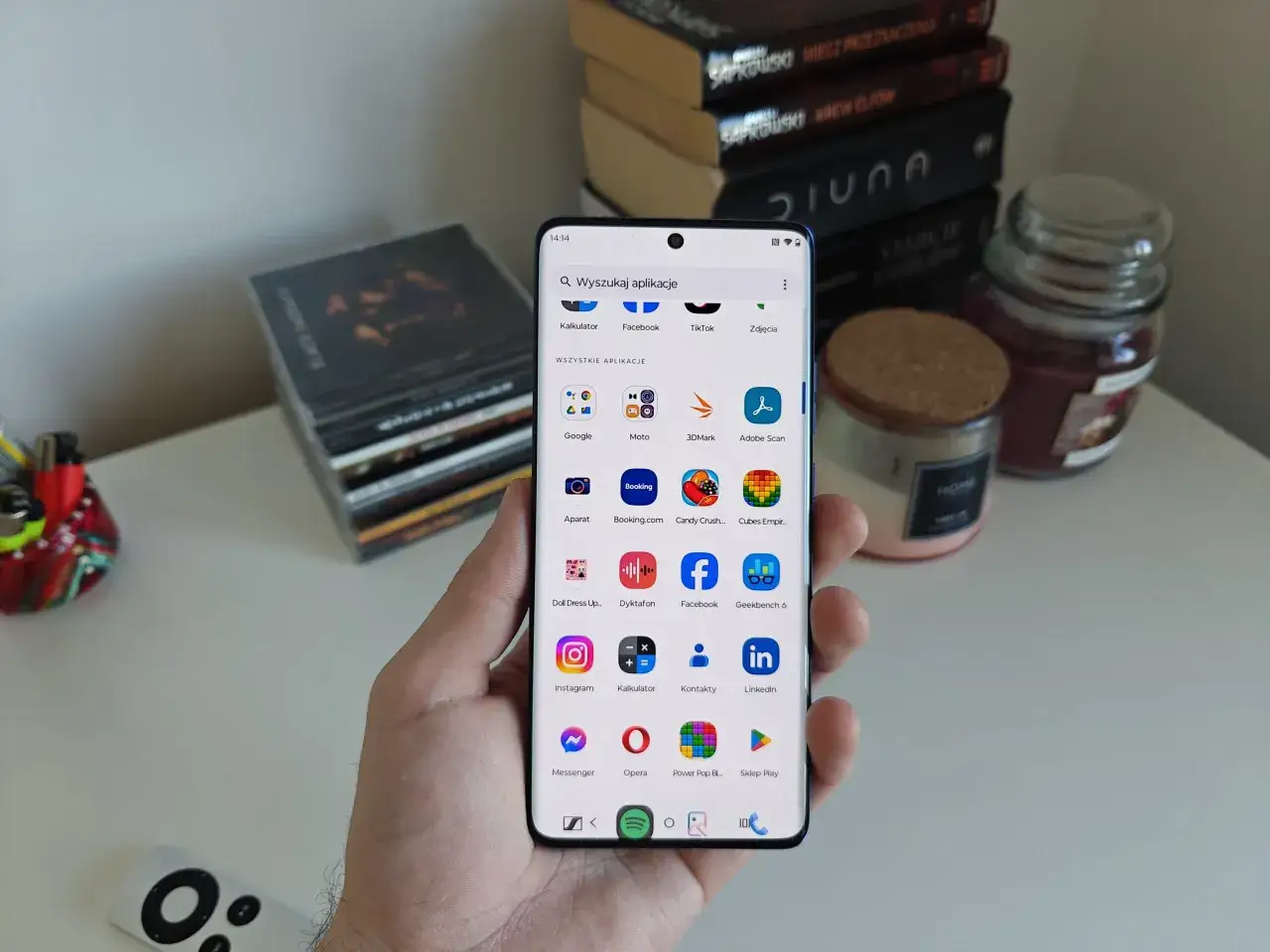 Dłoń trzyma telefon z czystym Androidem, wyświetlający ekran z aplikacjami. W tle stos książek i świece.