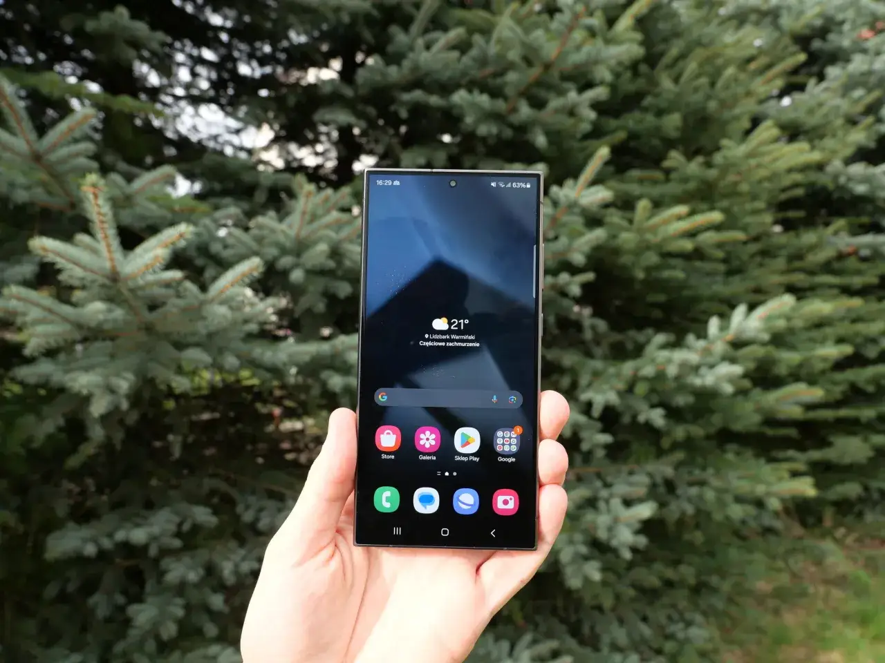Dłoń trzyma smartfon z interfejsem One UI, na tle iglastego drzewa. Widoczna jest pogoda i ikony aplikacji.