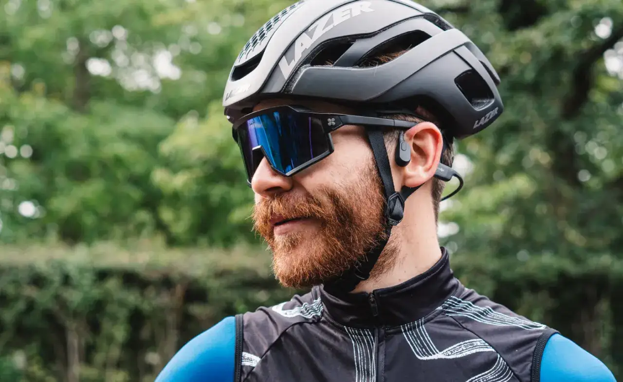 Rowerzysta w kasku i okularach z niebieskimi szkłami, z małymi słuchawkami w uszach. Idealne słuchawki na rower, by słyszeć otoczenie i muzykę.
