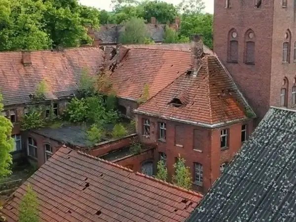 Opuszczony **szpital psychiatryczny** z rozpadającymi się dachami i zarośniętymi dziedzińcami, idealne miejsce na **horror o szpitalu psychiatrycznym**.