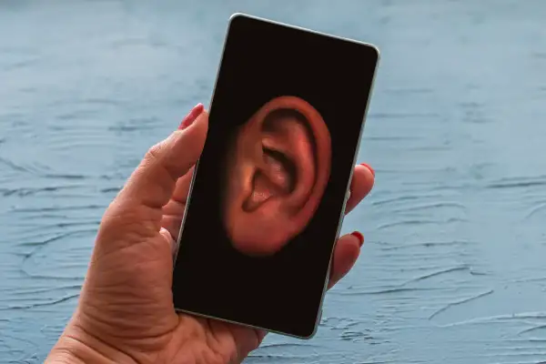 Dłoń trzyma smartfon z uchem na ekranie. Czy Twój telefon jest na podsłuchu? Dowiedz się, jak sprawdzić.