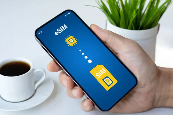 Dłoń trzyma smartfon z ekranem pokazującym grafikę symbolizującą play esim. Obok filiżanka kawy.