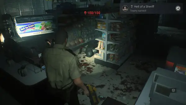 Policjant w sklepie, gdzie widać plamy krwi i półki z produktami. Osiągnięto trofea Resident Evil 2 "Hell of a Sheriff".